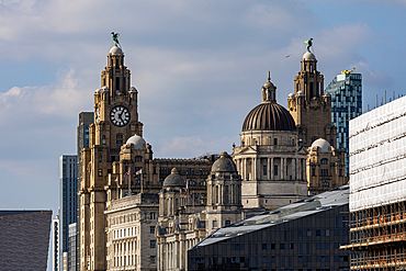 A detailed view of historic stone buildings, including a clock tower and domes, set against a cloudy sky. Modern buildings and scaffolding are also visible in Liverpool, UK.