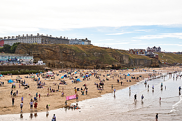 A wide, sandy beach is filled with people enjoying leisure activities under a partly cloudy sky. Buildings overlook the cliffside above the beach in Whitby, UK.
