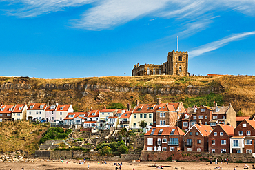 Coastal town with buildings nestled on a hillside beneath a prominent church structure. A sandy beach with scattered people lies in the foreground in Whitby, UK.