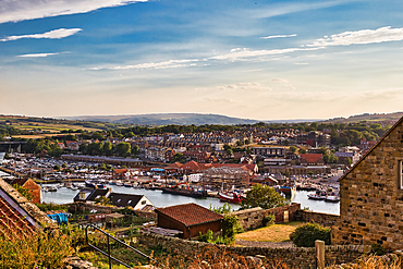 An elevated view captures a coastal town with a harbor filled with boats, nestled against rolling hills under a bright, partly cloudy sky in Whitby, UK.