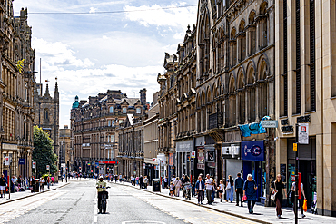 A wide street lined with historic stone buildings, featuring architectural details, shops, pedestrians, and a solitary cyclist in Newcastle upon Tyne, UK.