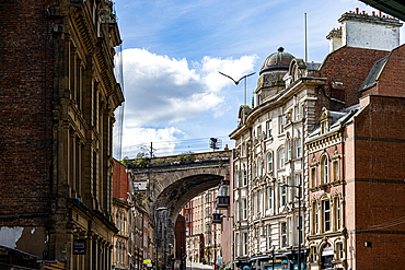 A view down a street lined with historic brick buildings, featuring an arched railway bridge in the distance under a partly cloudy blue sky in Newcastle upon Tyne, UK.