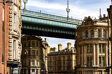 An urban scene featuring a large, teal-colored bridge spanning over ornate, stone buildings under a partly cloudy sky in Newcastle upon Tyne, UK.