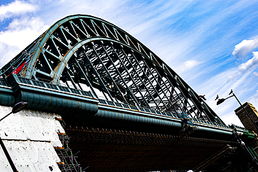 A low-angle view of a steel arch bridge with a complex lattice structure against a bright blue sky with wispy clouds in Newcastle upon Tyne, UK.