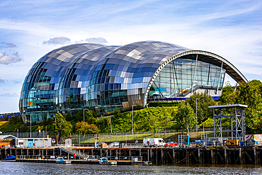 A modern building with a distinctive curved, glass and metal facade, sits on a green slope beside a calm body of water in Newcastle upon Tyne, UK.