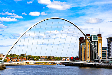 A white arched footbridge spans a river, with a cityscape and modern buildings on either side. Bright blue sky with scattered clouds in Newcastle upon Tyne, UK.