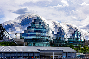 A modern, curved building with a distinctive tiled facade of silver, white, and blue panels, reflecting the cloudy sky in Newcastle upon Tyne, UK.