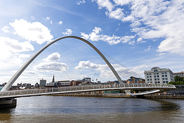 A modern white arch bridge spans a river with a cityscape and cloudy blue sky in Newcastle upon Tyne, UK.