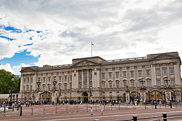 A wide shot of a grand palace with a flag flying from its roof, set against a cloudy sky. People are gathered in front of the gates in London, UK.