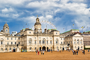 A grand, pale stone building with classical architecture dominates the frame, featuring a prominent clock tower. In the background, a large Ferris wheel is visible against a dramatic, cloudy sky.
