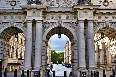 Grand stone arches frame a view of a street lined with classical buildings and a distant statue under a cloudy sky in London, UK.