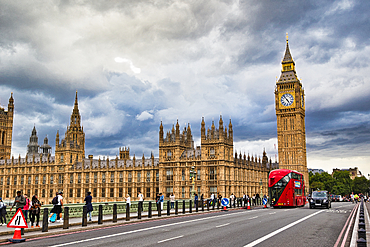 The Houses of Parliament and Big Ben tower are viewed from a bridge, with a red double-decker bus and black taxi on the road under a dramatic cloudy sky in London, UK.