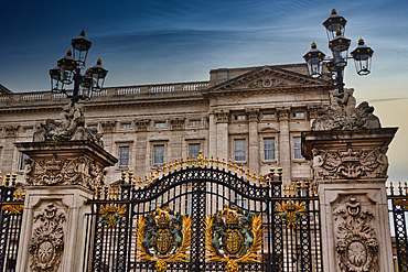 A grand black and gold gate with elaborate sculptures and ornate lamps in front of a pale stone building under a cloudy blue sky in London, UK.