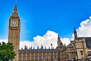 A wide shot captures the iconic Big Ben clock tower and the Houses of Parliament under a bright blue sky with scattered clouds in London, UK.