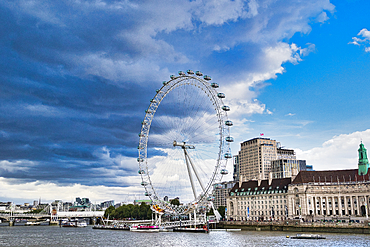 A large Ferris wheel stands prominently on the bank of a river, with modern and historical buildings lining the opposite shore under a dramatic sky.