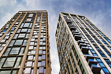 Two modern, high-rise apartment buildings with numerous windows and balconies, viewed from a low angle against a cloudy sky in London, UK.