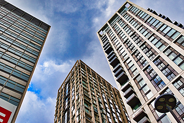 Three tall buildings with many windows reach towards a cloudy blue sky. The composition is a low-angle shot, emphasizing their height in London, UK.