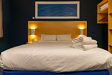 A hotel room with a king-sized bed, neatly made with white linens, two pillows, and a stack of white towels. Warm lighting emanates from wall sconces and illuminates the wooden headboard.