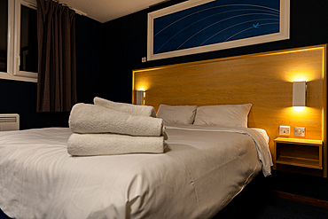 A hotel room features a made bed with white linens and stacked white towels, a wooden headboard with bedside lamps, and artwork on a dark blue wall.