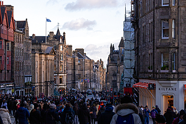 A crowded street scene of historic stone buildings, with people shopping and walking under a cloudy sky. A Scottish flag flies high in Edinburgh, Scotland.