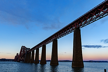 A low-angle view of a large red railway bridge spanning a body of water under a twilight sky.