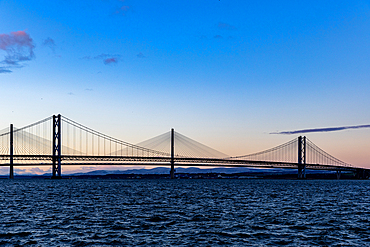 A wide suspension bridge spans across a dark, choppy body of water under a gradient sky of blues and soft oranges, with distant hills silhouetted.