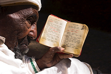 Old man wearing traditional gabi (white shawl) sitting in the sun reading holy bible, at church of Bet Medhane Alem (Saviour of the World), Lalibela, World Heritage Site, Ethiopia, Africa