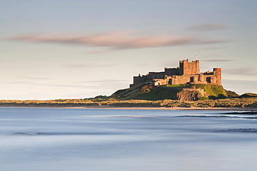 Bamburgh Castle bathed in golden evening light overlooking Bamburgh Bay with the sea filling the foreground, Northumberland, England, United Kingdom, Europe