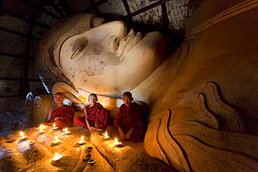 Three novice Buddhist monks sitting by a large reclining Buddha in the light of candles, near Shwesandaw Paya, Bagan, Myanmar (Burma), Asia