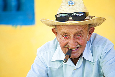 Portrait of old man wearing straw hat and smoking cigar, posing against a yellow wall for tourist pesos, Trinidad, Cuba, West Indies, Central America