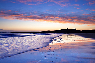 Looking across Embleton Bay at sunrise towards the silhouetted ruins of Dunstanburgh Castle in the distance and the vivid colours in the sky reflecting in the sea and wet sand, Embleton, near Alnwick, Northumberland, England, United Kingdom