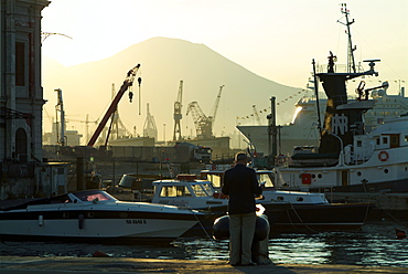 Italy, Campania, Naples, port, man reading newspaper with docks and Vesuvius behind