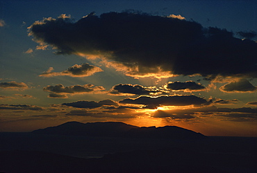 Sun behind dark clouds over hills and coast