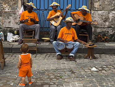 Street band wearing orange shirts playing music on the pavement watched by toddler wearing orange clothes, Habana Vieja (Old Havana), Havana, Cuba, West Indies, Central America