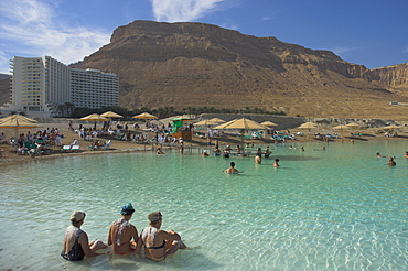 Three women sitting in the sea and Hyatt hotel and desert cliffs in background, Ein Bokek Hotel Resort, Dead Sea, Israel, Middle East 