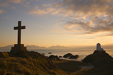 Stone Cross and old lighthouse (Twr Mawr) in silhouette at sunset on rocky tip of Llanddwyn Island National Nature Reserve, Newborough, Anglesey, North Wales, United Kingdom, Europe