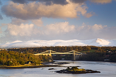 Menai Suspension Bridge built by Thomas Telford in 1826, across the Menai Strait with snow on mountains of Snowdonia in winter evening light, Anglesey, North Wales, United Kingdom, Europe