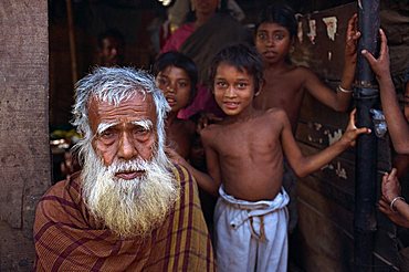 Head and shoulders portrait of an old Bangladeshi (Bengali) man with a white beard, and children beyond, looking at the camera, in the slums of Dhaka (Dacca), Bangladesh, Asia