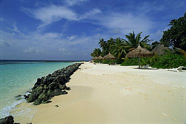 Tropical beach with thatched umbrellas and line of rocks on the edge of the sea at Nakatchafushi in the Maldive Islands, Indian Ocean, Asia