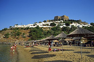 The beach below the white houses and acropolis of Lindos Town, Rhodes, Dodecanese Islands, Greek Islands, Greece, Europe