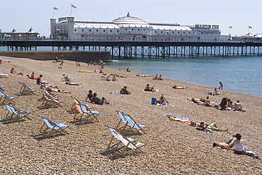 Deck chairs and sunbathers on the pebble beach in view of Brighton Pier, Brighton, Sussex, England, United Kingdom, Europe