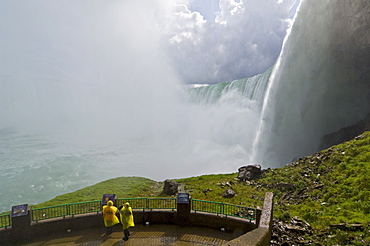 Two tourists in yellow raincoats in the spray of the Horseshoe Falls waterfall whilst on the Journey under the Falls tour at Niagara Falls, Ontario, Canada, North America