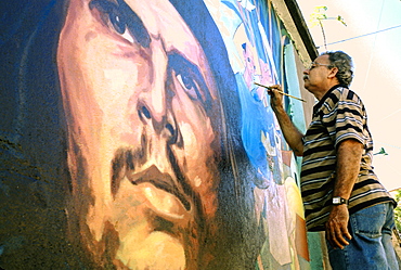 An artist painting a wall mural featuring a portrait of the revolutionary hero Che Guevara on a street in Baracoa, Cuba