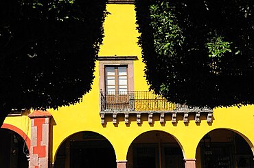 Mexico, Bajio, San Miguel de Allende, El Jardin Detail of yellow painted facade of colonial mansion with French window and balcony part framed by trees in foreground.