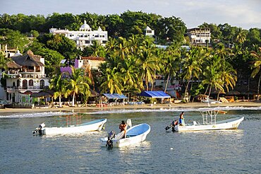 Mexico, Oaxaca, Puerto Escondido, Fishing boats in bay at Playa Marinero lined with cafes bars and other buildings amongst palm trees.