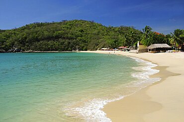 Mexico, Oaxaca, Huatulco, Sandy beach at Bahia Santa Cruz lined by restaurants and other buildings with tree covered coastline beyond.