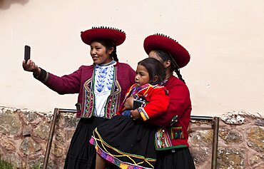 Peru, Chinchero, Young woman in traditional dress using a cell phone to take a picture of herself and other woman with a child.