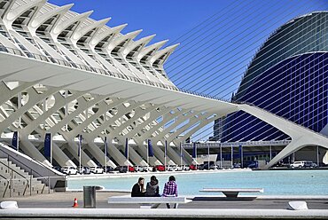 Spain, Valencia Province, Valencia, La Ciudad de las Artes y las Ciencias, City of Arts and Sciences, Principe Felipe Science Museum, General view of the building with girls on seat in the forground and row of Mercedes cars below the building.