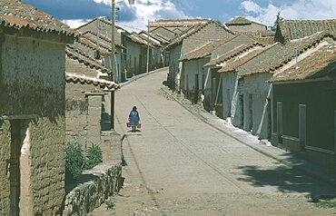 BOLIVIA Chuquisaca Tarabuco Town in the Altiplano or Bolivian Plateau.  Woman carrying two buckets walking along narrow paved street between houses with overhanging tiled rooftops.