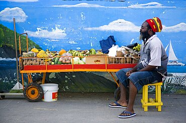 WEST INDIES St Vincent & The Grenadines Union Island Fruit and vegetable stall holder in front of wall painting in Clifton